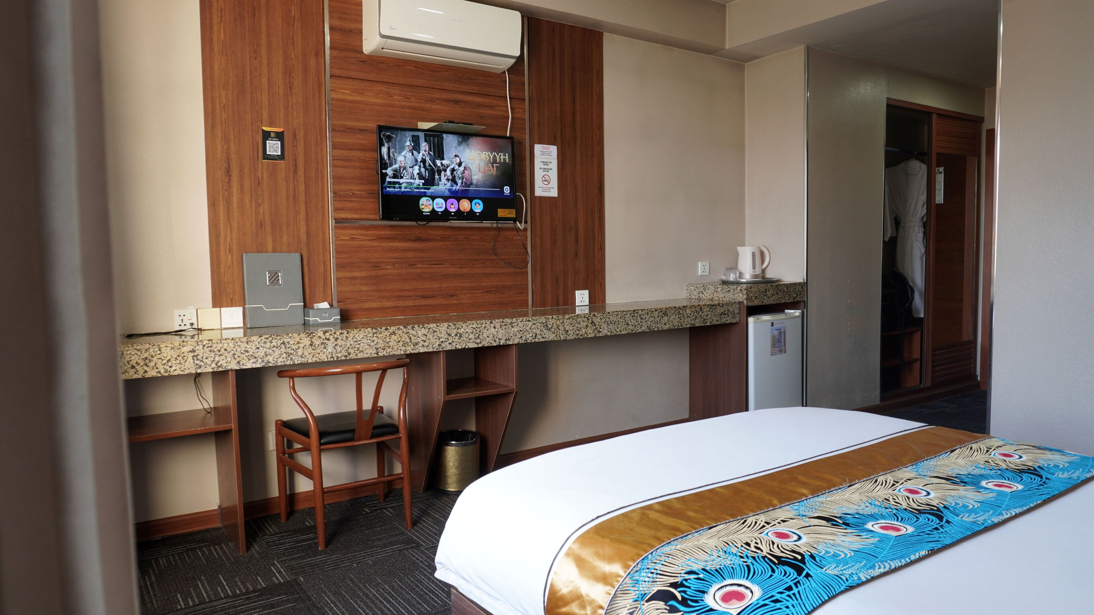 Habitación de hotel con cama, escritorio con silla, televisor montado en la pared y un refrigerador pequeño.
