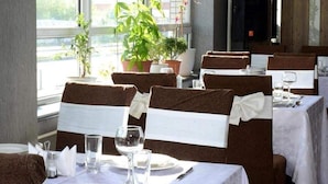 Un restaurante con mesas dispuestas para comer, sillas con cintas blancas y plantas en macetas cerca de las ventanas.