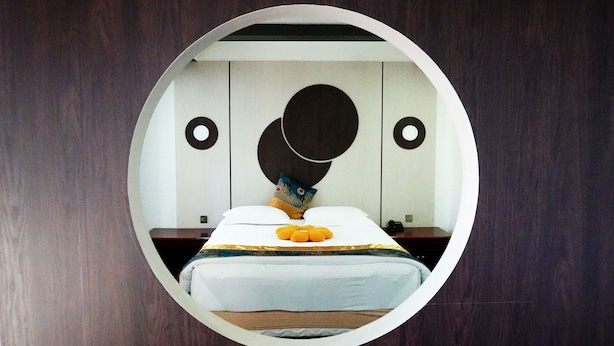 Un espejo circular refleja un dormitorio con cama, almohadas y una mesita de noche.