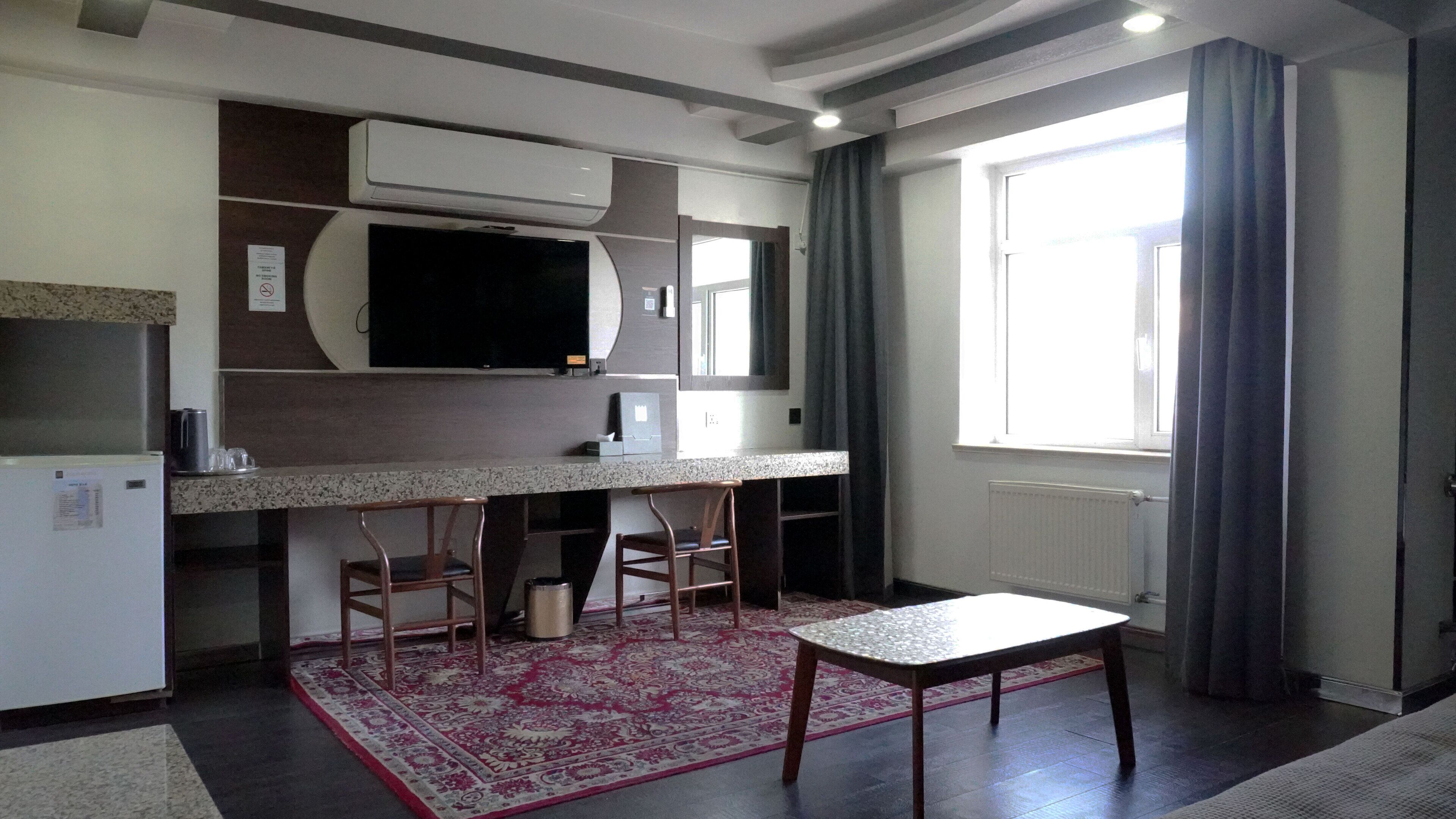 Cocina compacta con microondas integrado, un refrigerador pequeño y un televisor de pantalla plana. Hay un taburete de bar y una mesa pequeña frente a la encimera. La habitación tiene un ventanal con cortinas y un tapete estampado en el suelo.