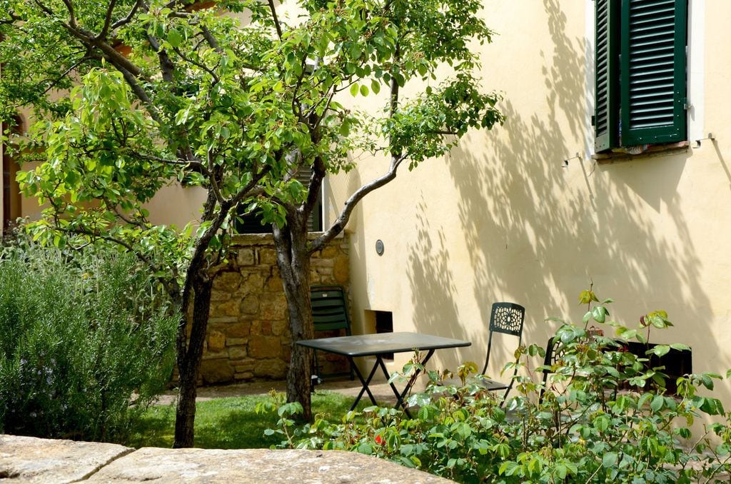 A courtyard with a stone wall, a table, and chairs, surrounded by greenery and a building with a window and shutters.