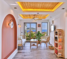 Una sala de estar moderna con techo de madera, puerta de vidrio que da a un balcón con vista al mar, muebles de mimbre y una estantería.