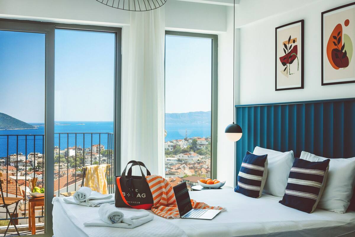 A hotel room with a bed, a laptop, a handbag, and a bowl of fruit. There are two large windows with a view of the sea and buildings.