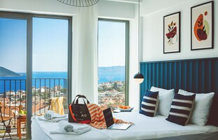 Habitación de hotel con una cama, una computadora portátil, un bolso y un frutero. Hay dos ventanas grandes con vista al mar y edificios.