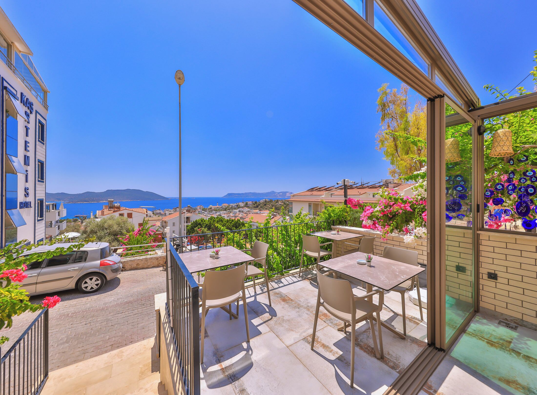A hotel balcony with outdoor seating, a clear view of the sea, and a cityscape in the background.