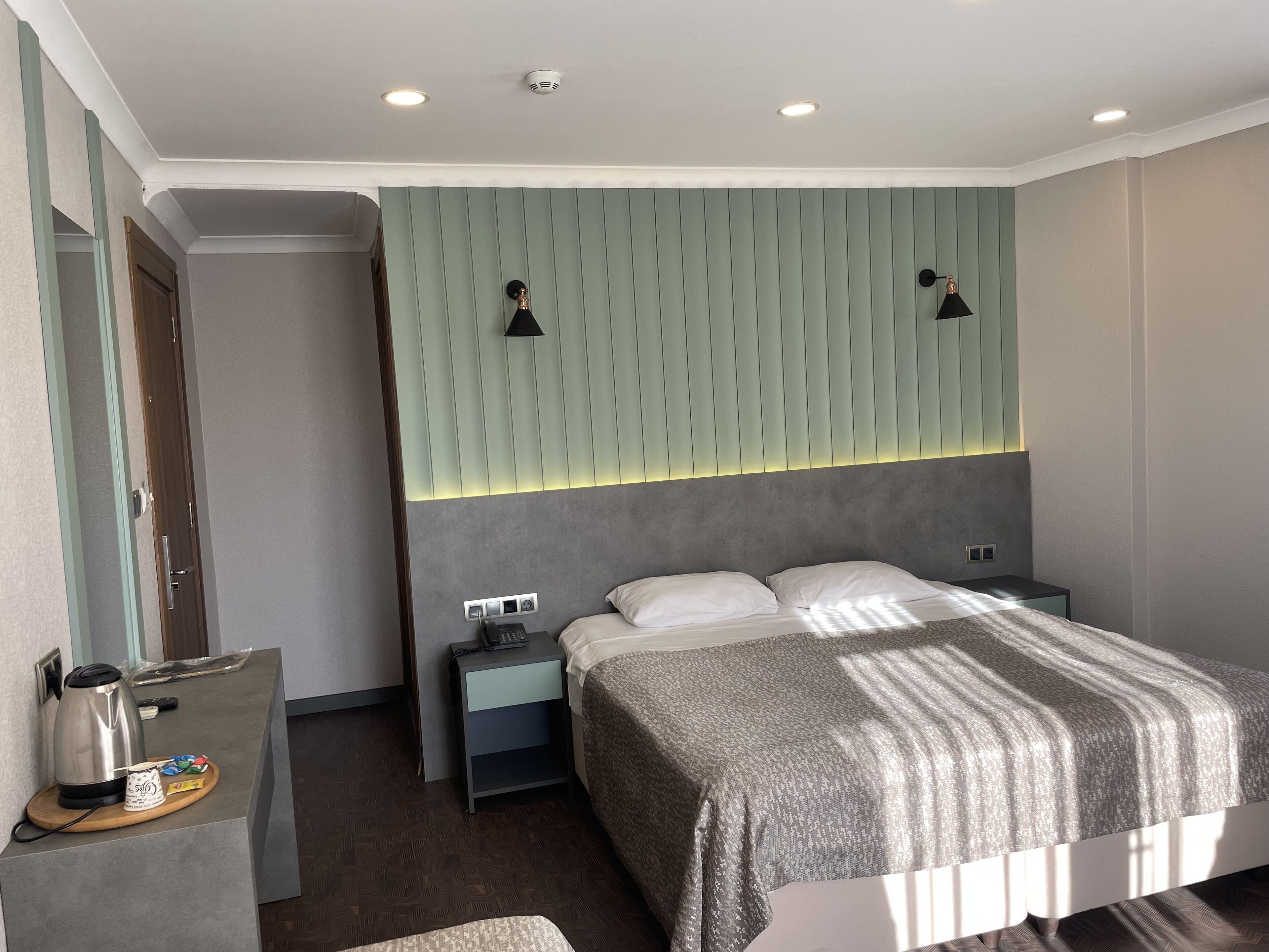 Une chambre d’hôtel moderne équipée d’un lit, d’une table de chevet et d’un petit bureau avec une bouilloire et du café.