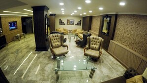 A lobby with a glass-top coffee table surrounded by armchairs, a sofa, and a TV screen displaying a news channel. - Keles Hotel (Rize)