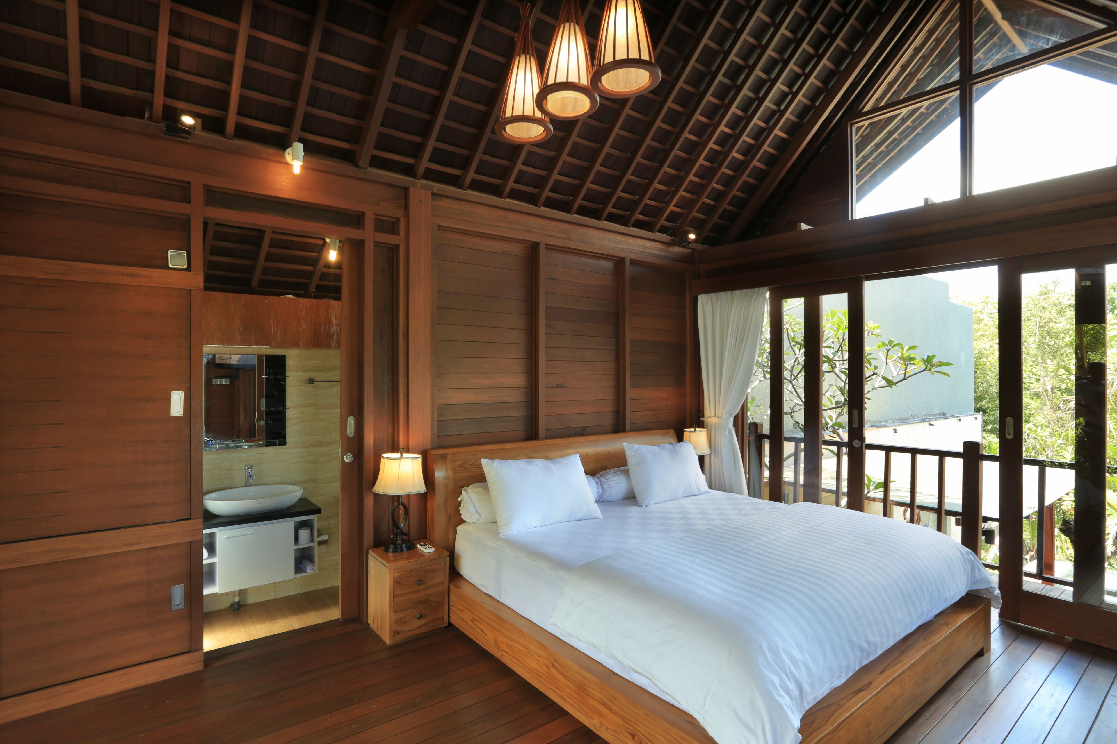 Une chambre à coucher en bois avec un grand lit, des lampes de chevet, une table de chevet en bois, un lavabo intégré et une grande fenêtre donnant sur un paysage verdoyant.