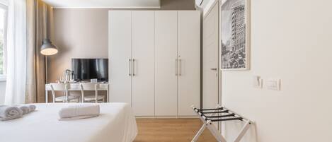 A hotel room with a bed, a desk with a TV, a wardrobe, and a drying rack.