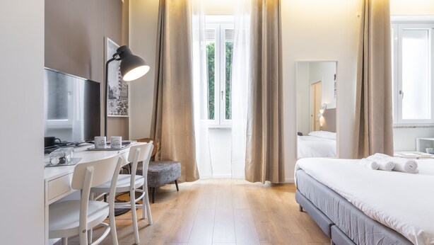 A hotel room with a bed, a desk with a computer, a chair, a window with curtains, and a bedside table. - Dome (Cagliari)