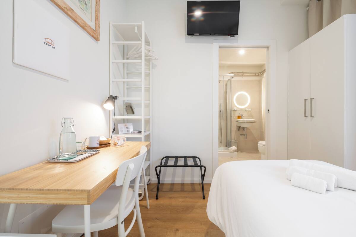 A hotel room with a wooden desk, white chairs, a bed with white linens, a TV mounted on the wall, and a bathroom visible through an open door.
