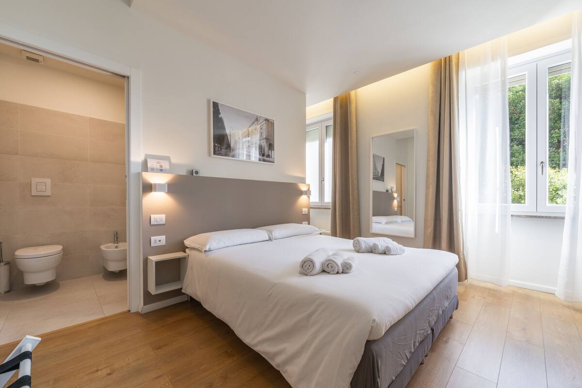 A modern hotel room with a large bed, bedside tables, a bathroom with a toilet and bidet, and a window with curtains.