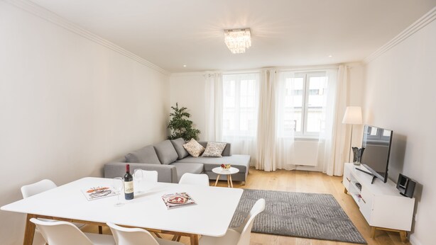 Una sala moderna con un sofá blanco, una mesa de comedor con sillas, un televisor sobre un mueble blanco y una araña.