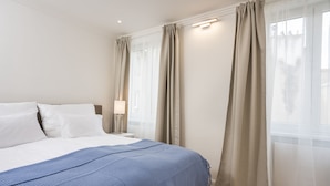 Una cama bien hecha con ropa de cama blanca y azul, una lámpara de noche y cortinas translúcidas en una ventana.