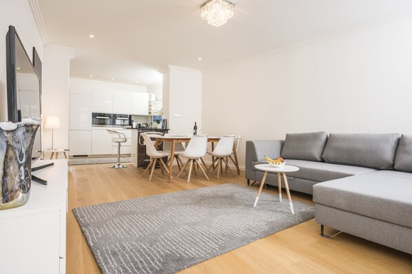 Uma sala de estar moderna com um sofá cinzento, uma mesa redonda pequena e uma área de jantar com mesa de madeira e cadeiras brancas.