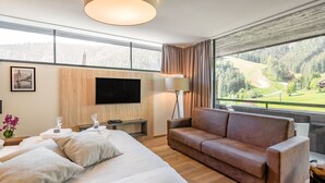Moderni hotellihuone, jossa on suuri sänky, nahkasohva, taulutelevisio ja näkymä vuoristomaisemaan.