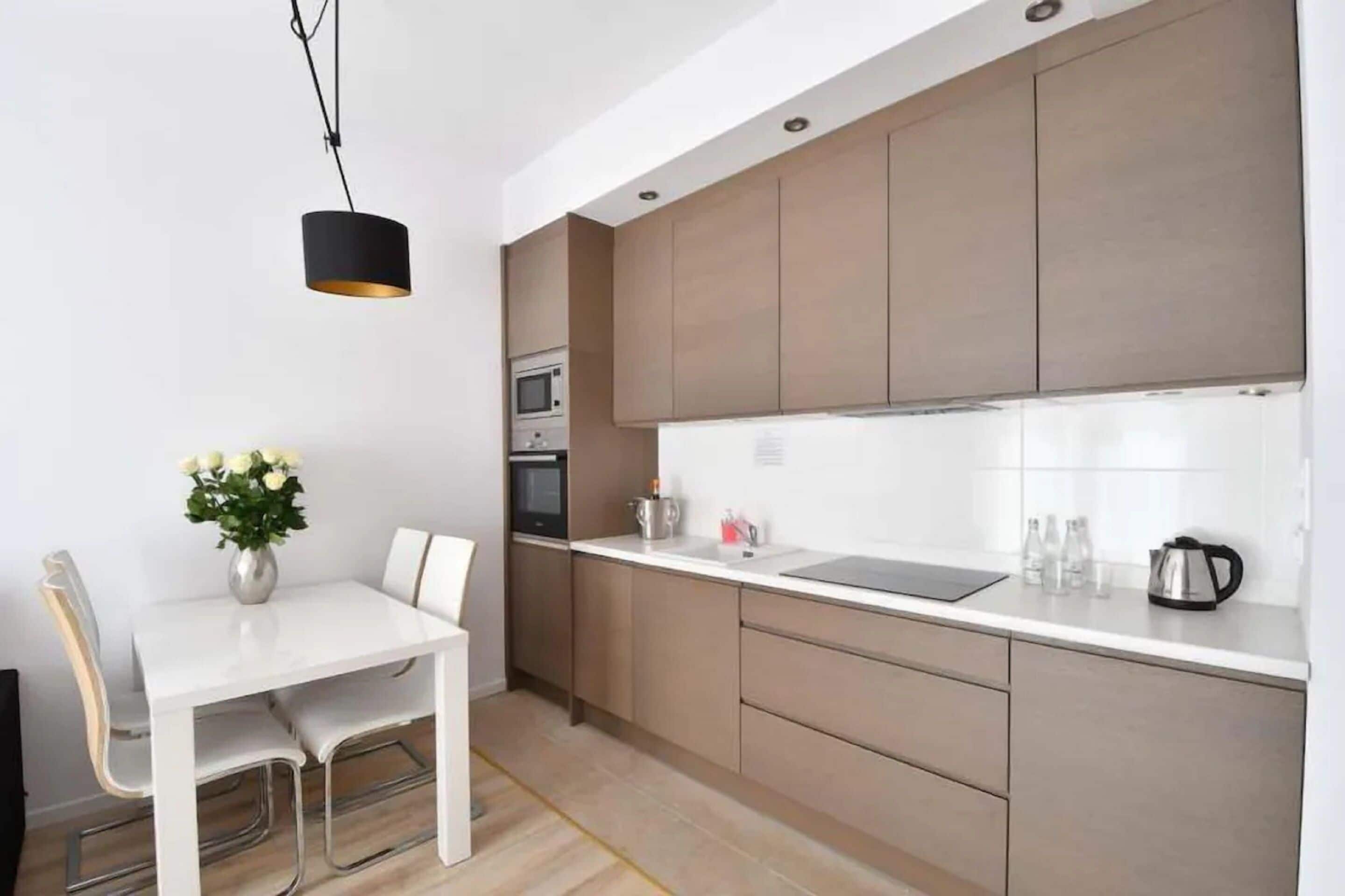 Une cuisine moderne avec des meubles blancs, un luminaire suspendu noir, un four à micro-ondes et un coin repas comprenant une table et des chaises blanches.