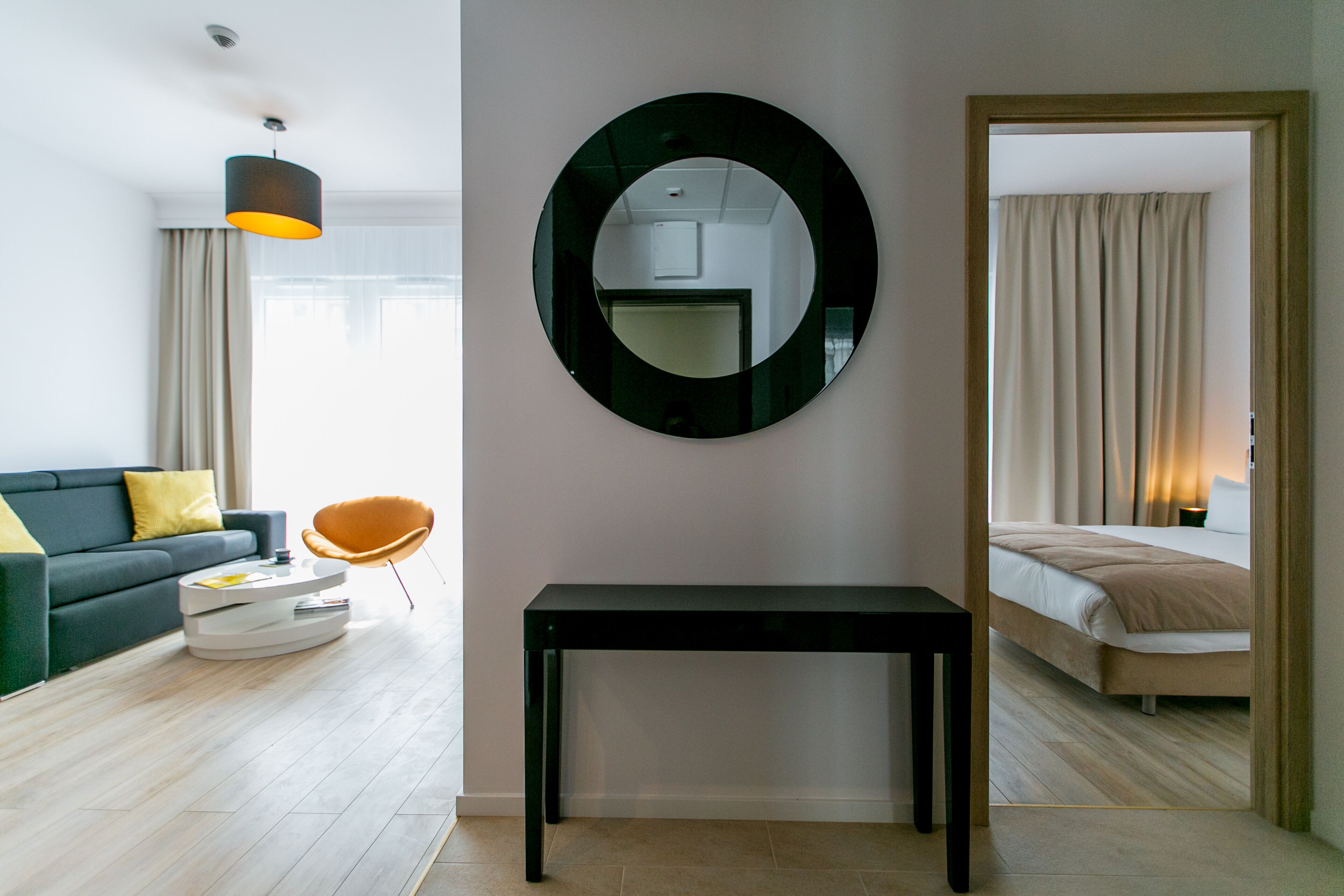 A modern hotel room with a round mirror, a black console table, a green sofa with yellow cushions, a white coffee table, and a bed with beige curtains.