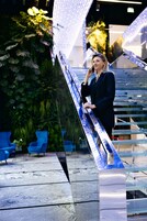 Una mujer está de pie en una escalera mecánica metálica en un espacio interior moderno, con sillas azules y follaje verde al fondo.