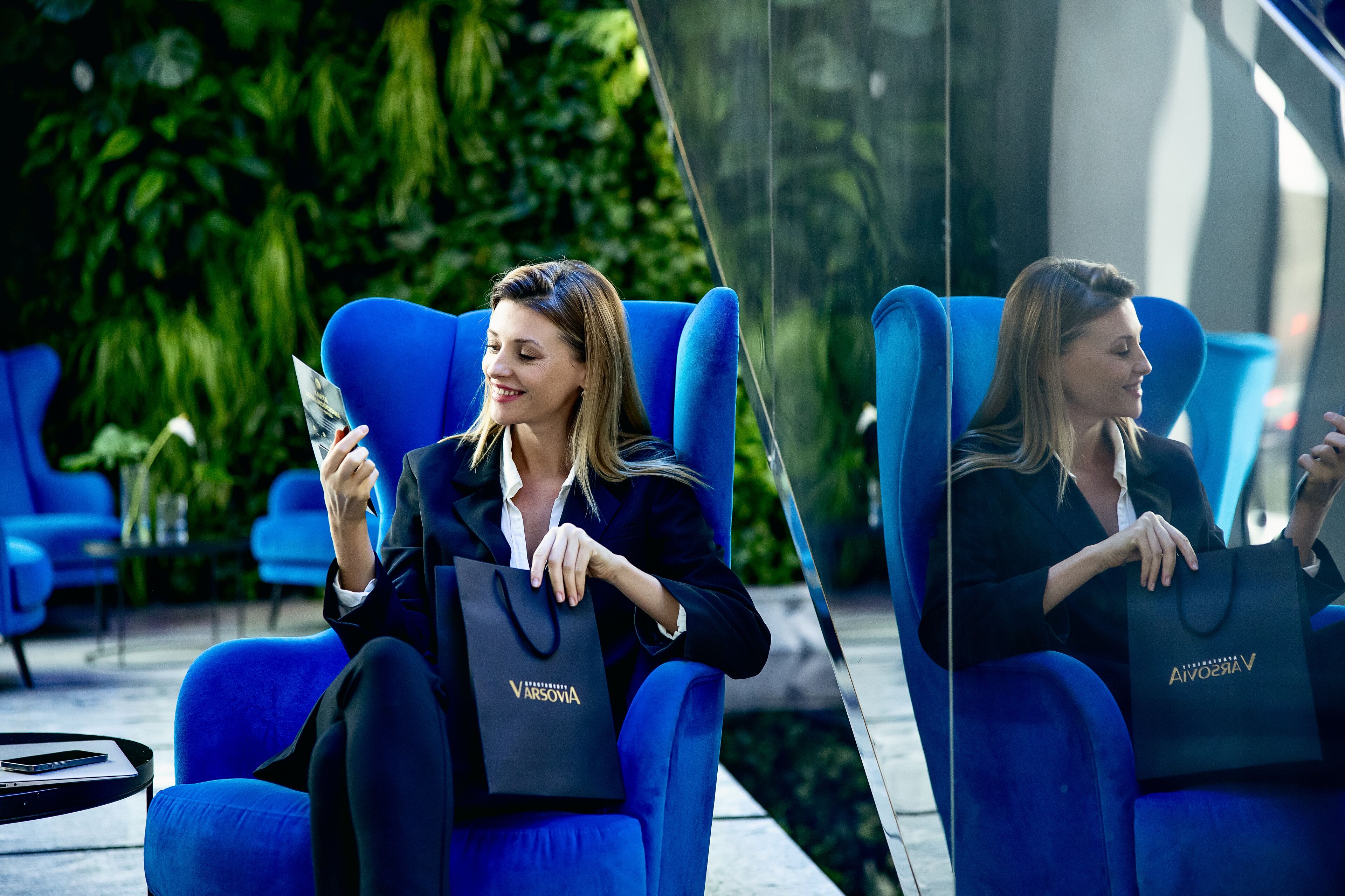 Une personne assise sur une chaise bleue, avec un sac noir sur lequel est inscrit le mot « VARSOVIE » et un téléphone intelligent.