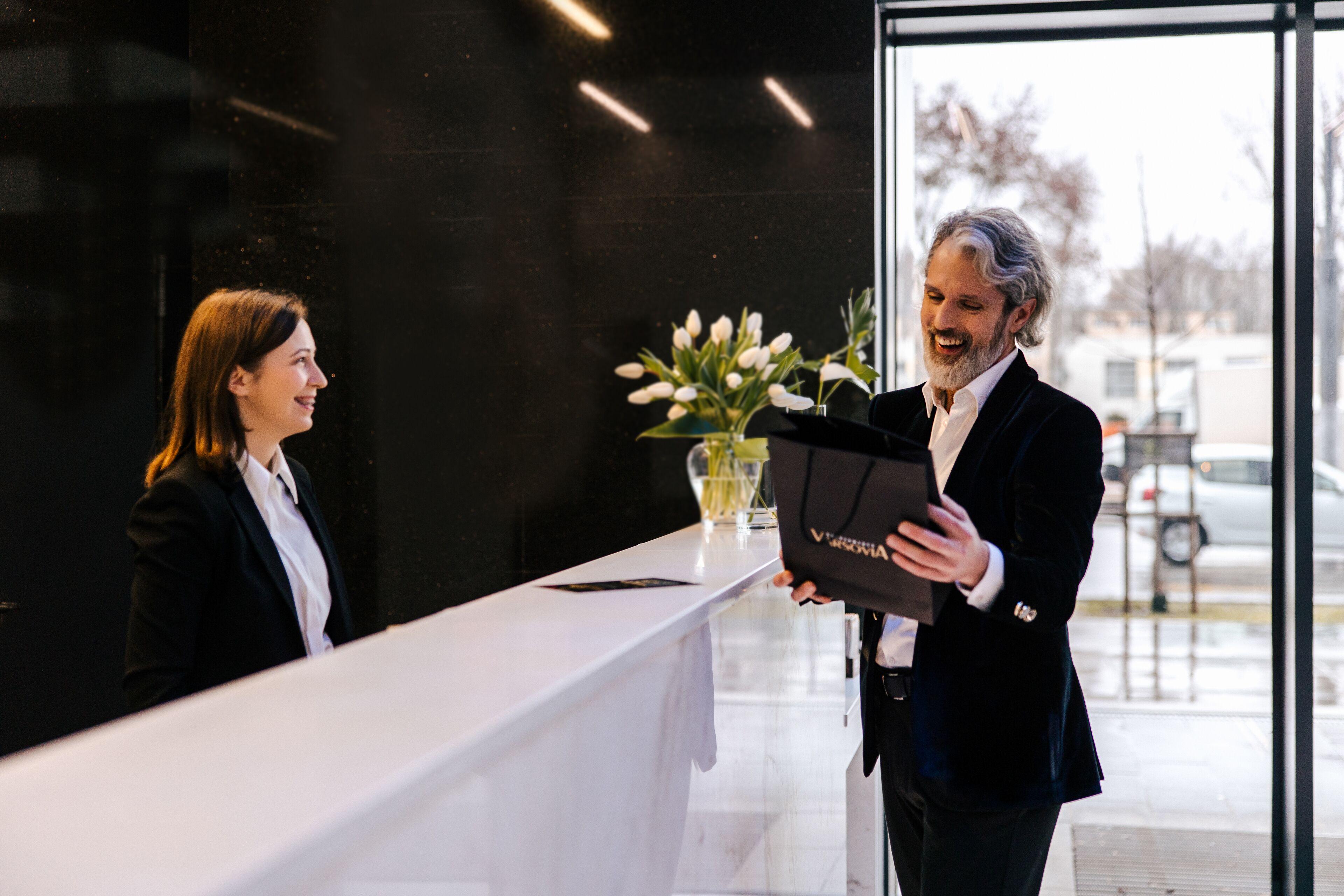 A reception area with a white reception desk, a vase with flowers, and a man holding a black folder.