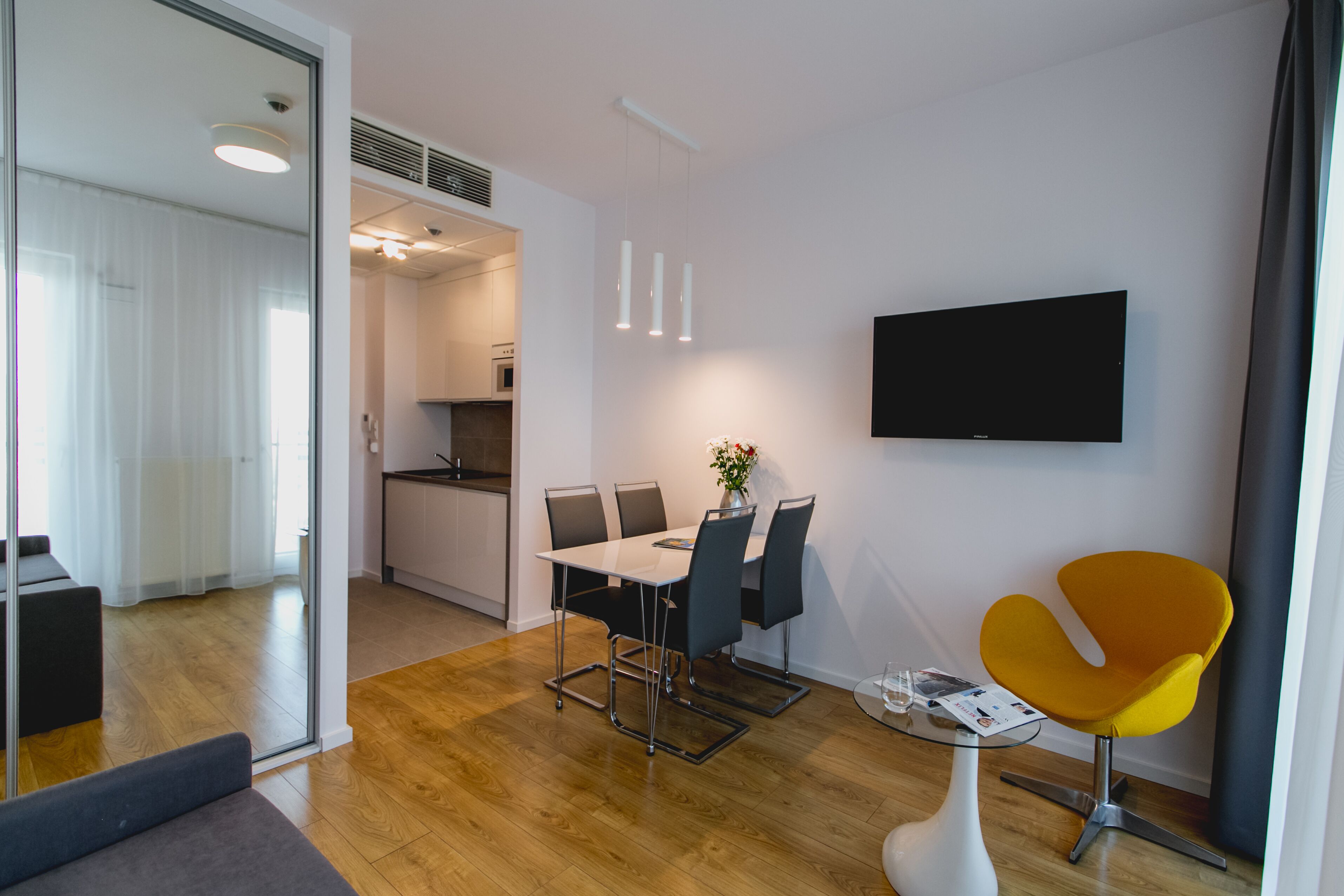 Una sala de estar moderna con comedor, una cocina al fondo y un televisor montado en la pared.