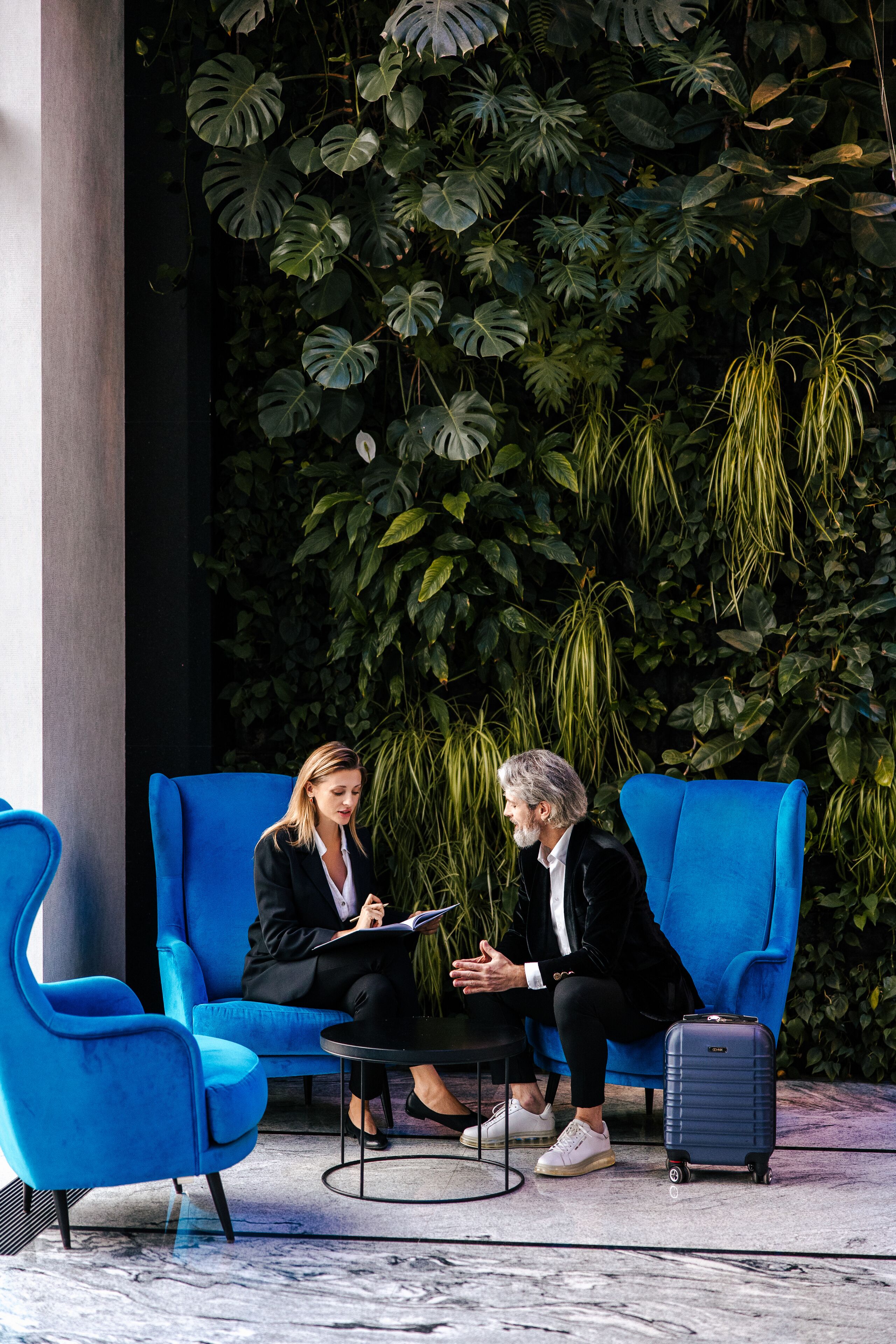 Two people seated on a blue sofa in an indoor setting with a backdrop of large green plants.