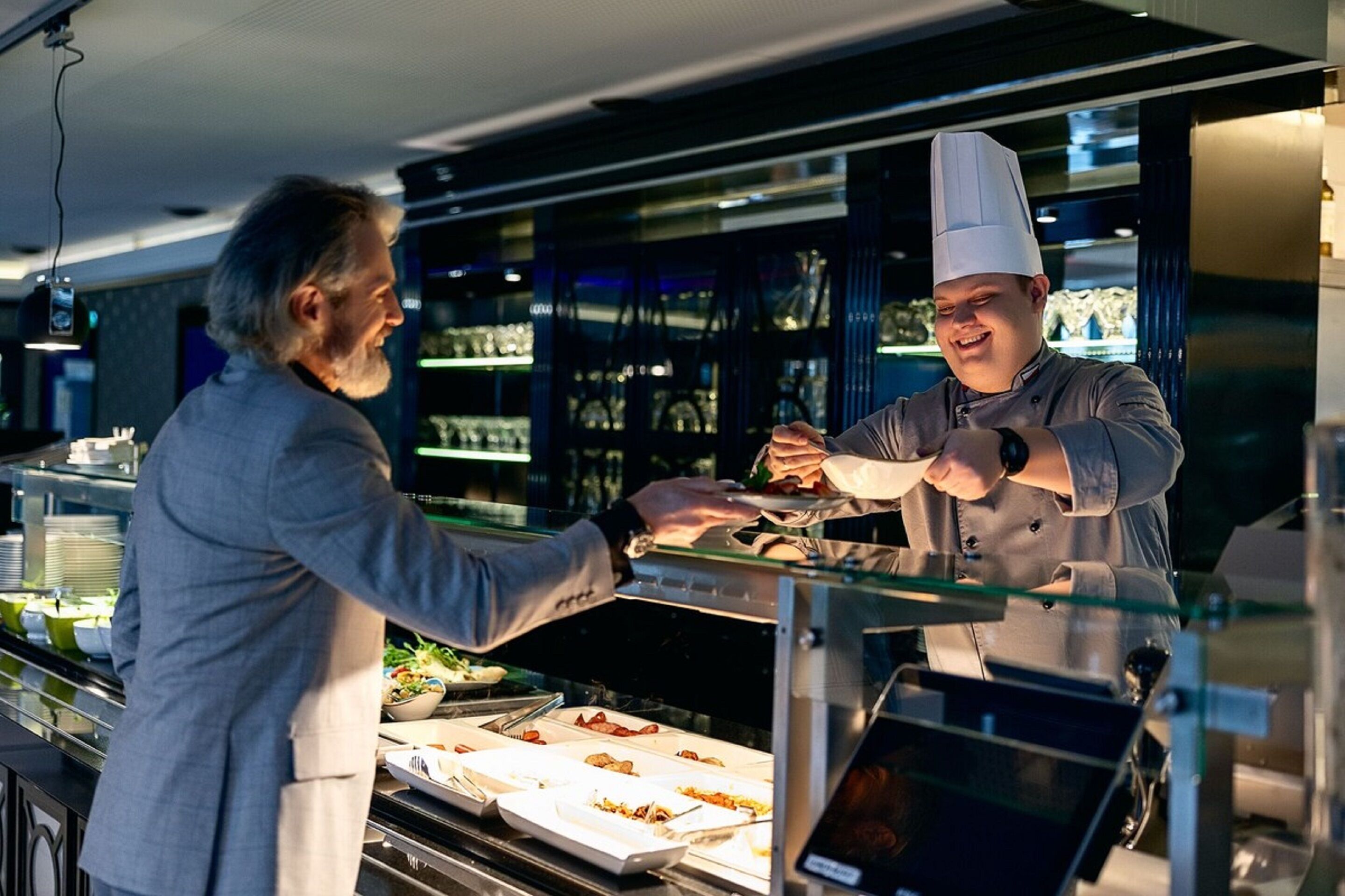 Zona de bufé con mampara de vidrio, bandejas de comida y pantalla digital con el menú.