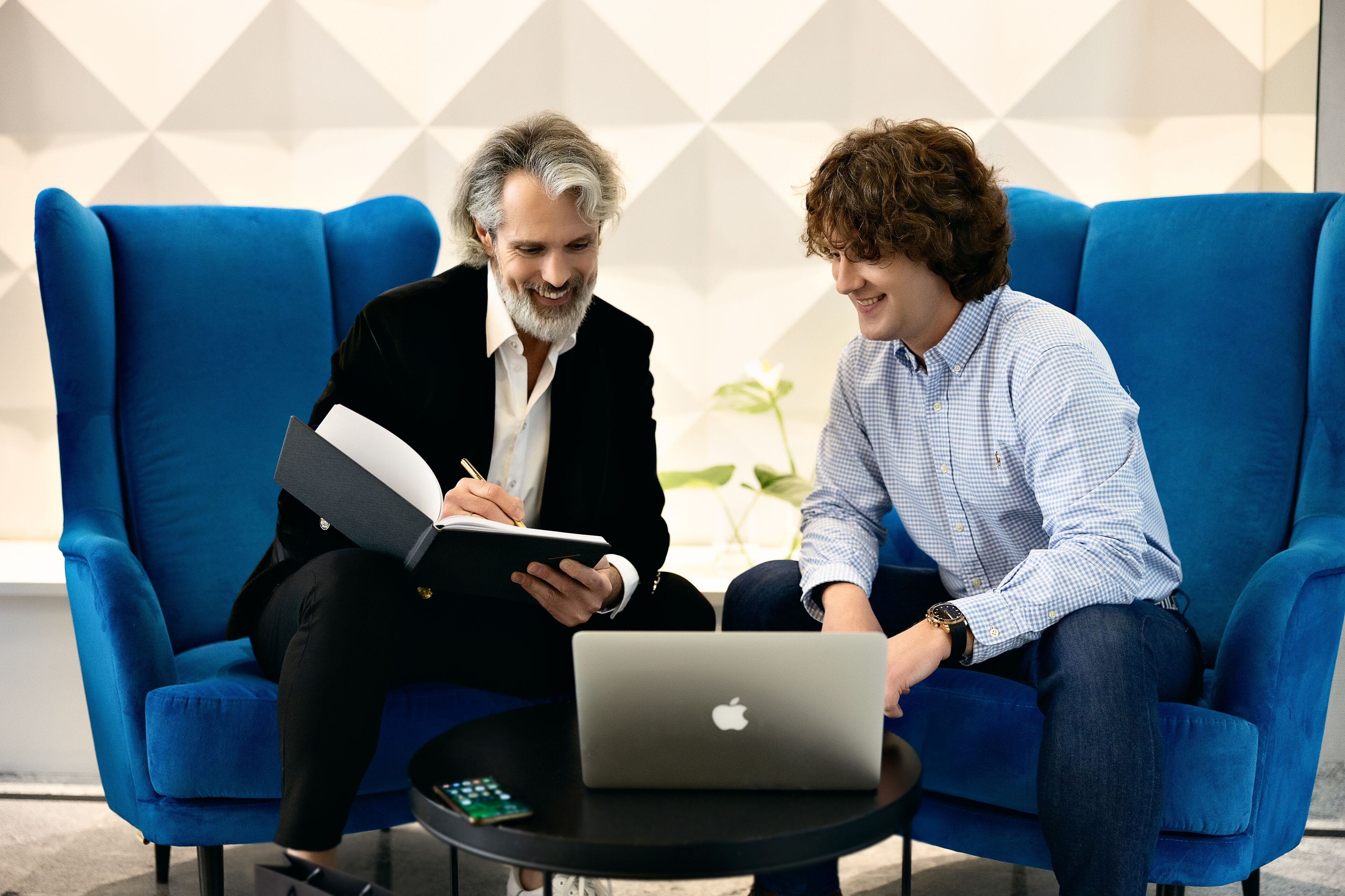Two people sitting on a blue couch with a laptop on a small table between them.
