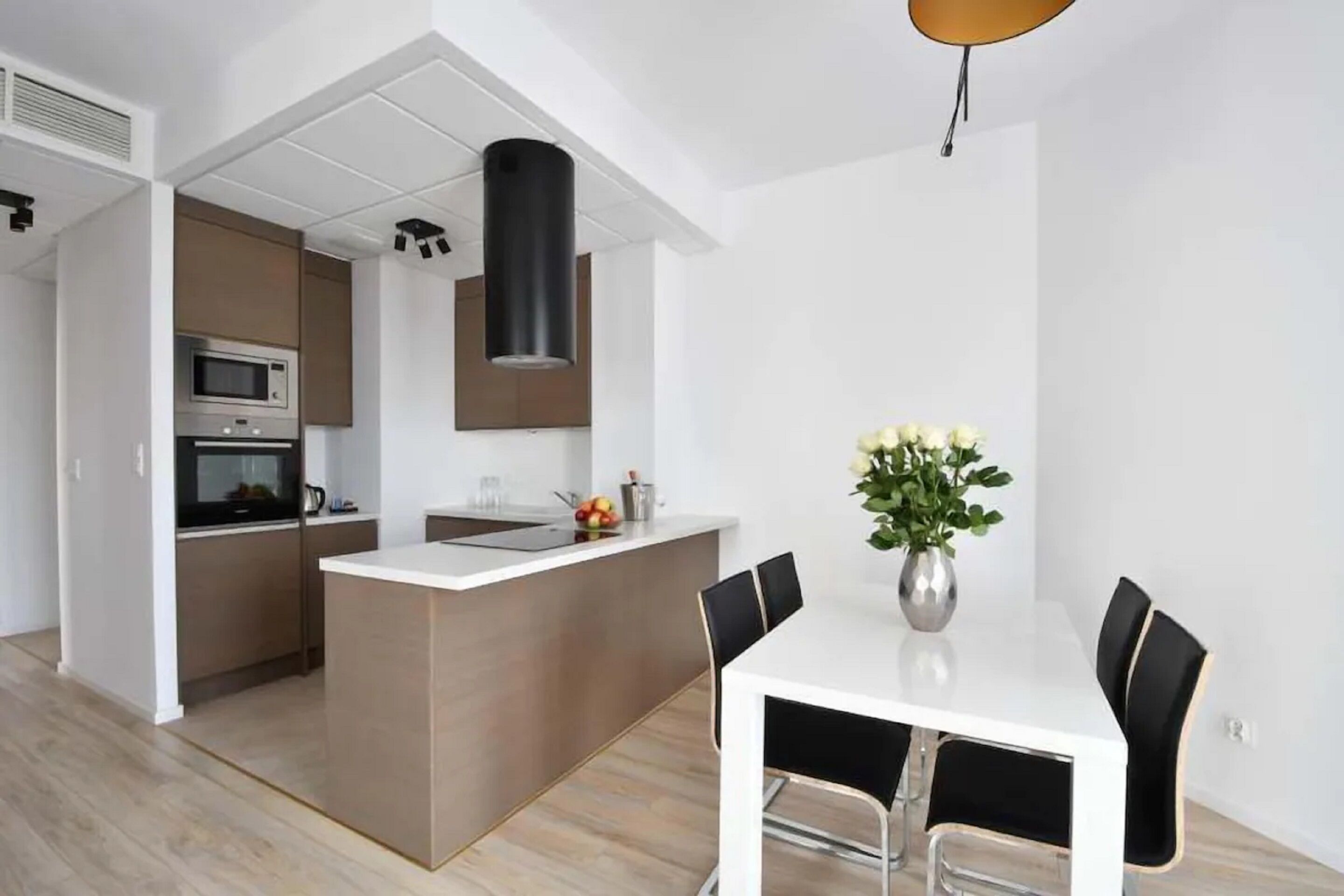 A modern kitchen with a central island, a dining area with a white table and black chairs, and a hanging light fixture.