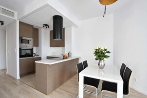 A modern kitchen with a central island, a dining area with a white table and black chairs, and a hanging light fixture.