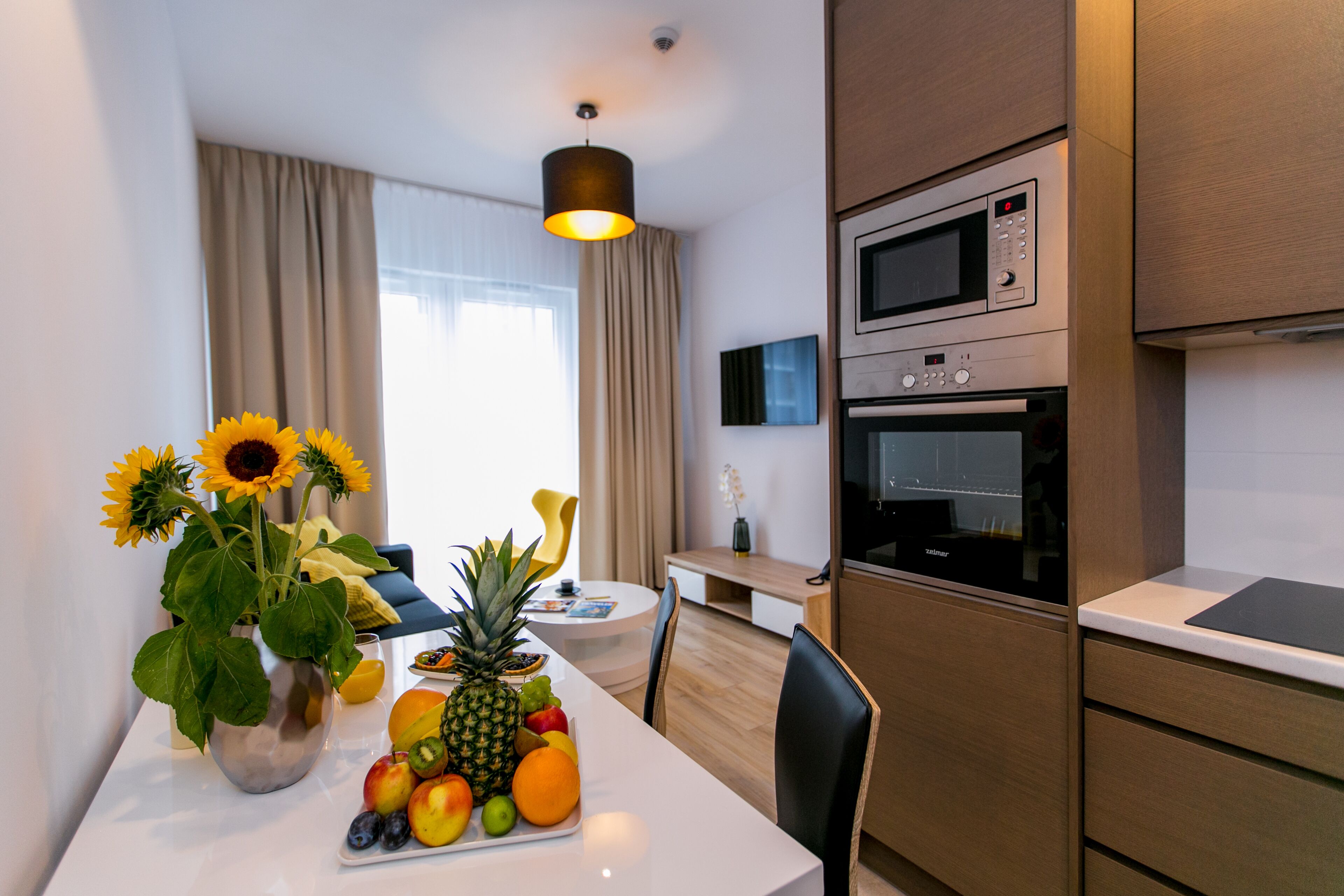 A modern kitchen with a microwave, oven, and a dining area with a fruit bowl and a vase of sunflowers.
