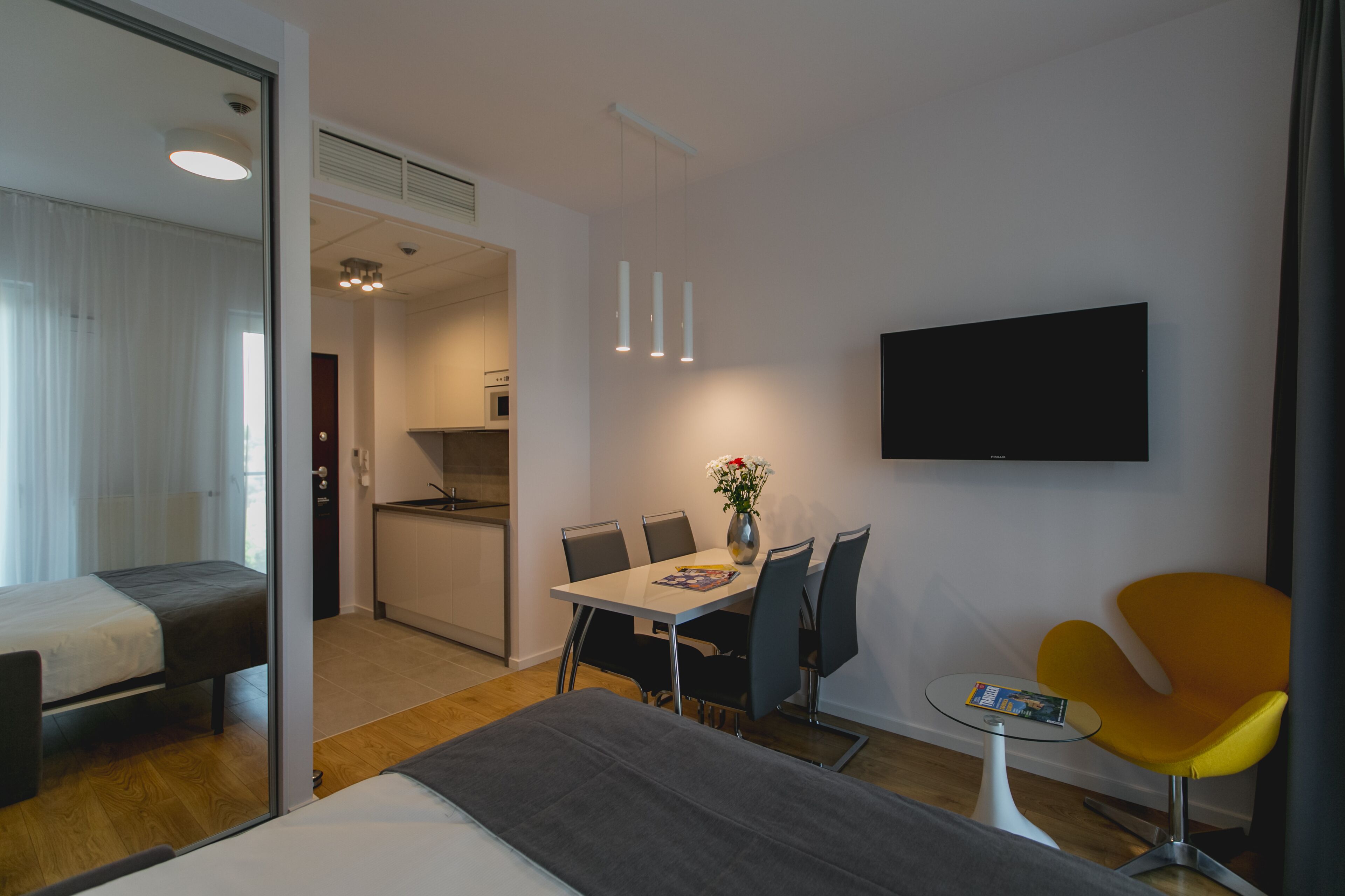 Ein modernes Hotelzimmer mit einem Bett, einem Essbereich mit Tisch und Stühlen, einem kleinen Tisch mit einer Zeitschrift, einem gelben Sessel und einem an der Wand befestigten Fernseher.