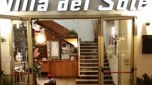 The entrance of Villa del Sole with a wooden deck, a staircase leading inside, and a reception area with a counter and plants.