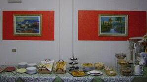 A room with two framed paintings on the walls, a table with various food items and plates, and a bed with a patterned bedspread.