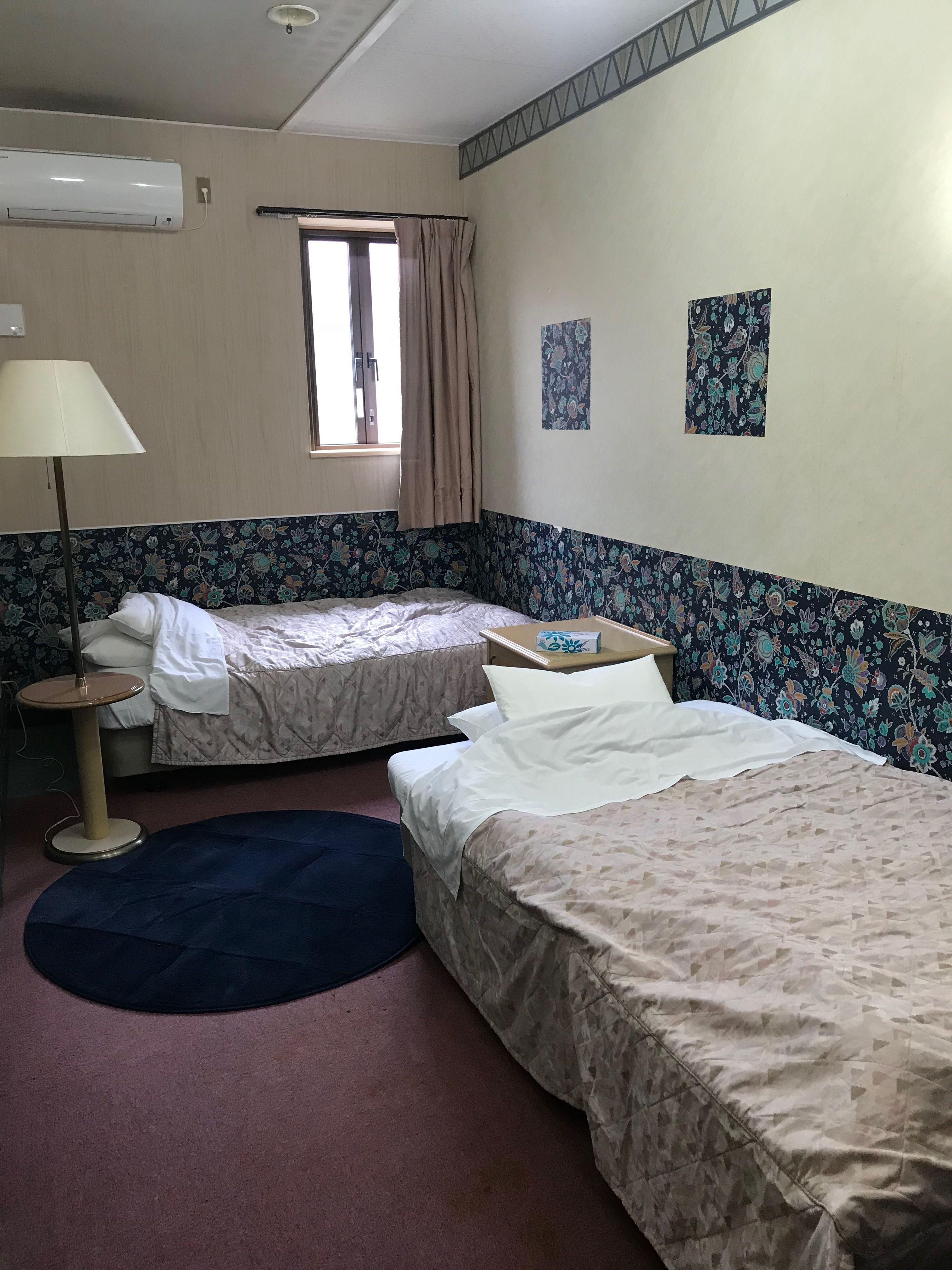 Habitación de hotel con dos camas, un escritorio, una lámpara, una ventana con cortinas y una pared con papel tapiz floral.