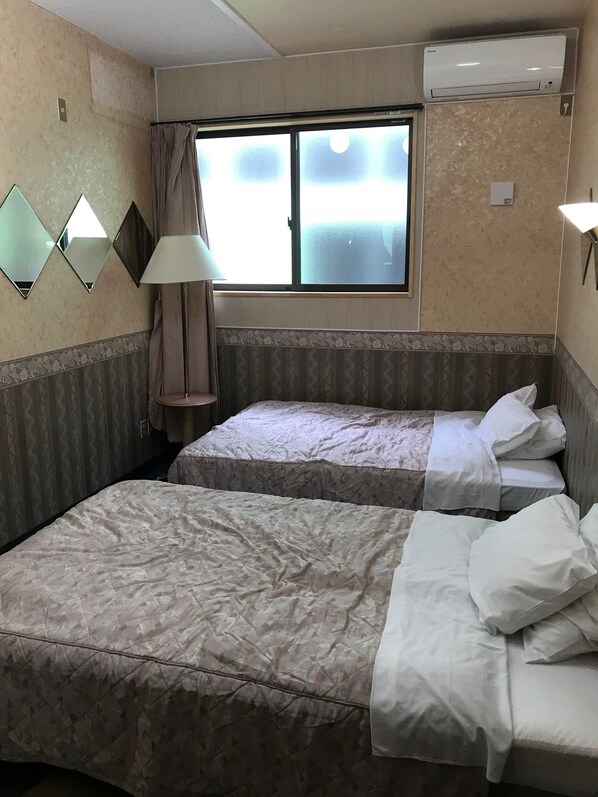 Habitación de hotel con dos camas, una ventana con cortinas, aire acondicionado instalado en la pared y una lámpara.