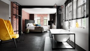 A modern hotel room with a large bed, a yellow armchair, a white bathtub, and a view of a brick wall through the window. - Aparthotel Mittermeiers Alter Ego (Rothenburg ob der Tauber)