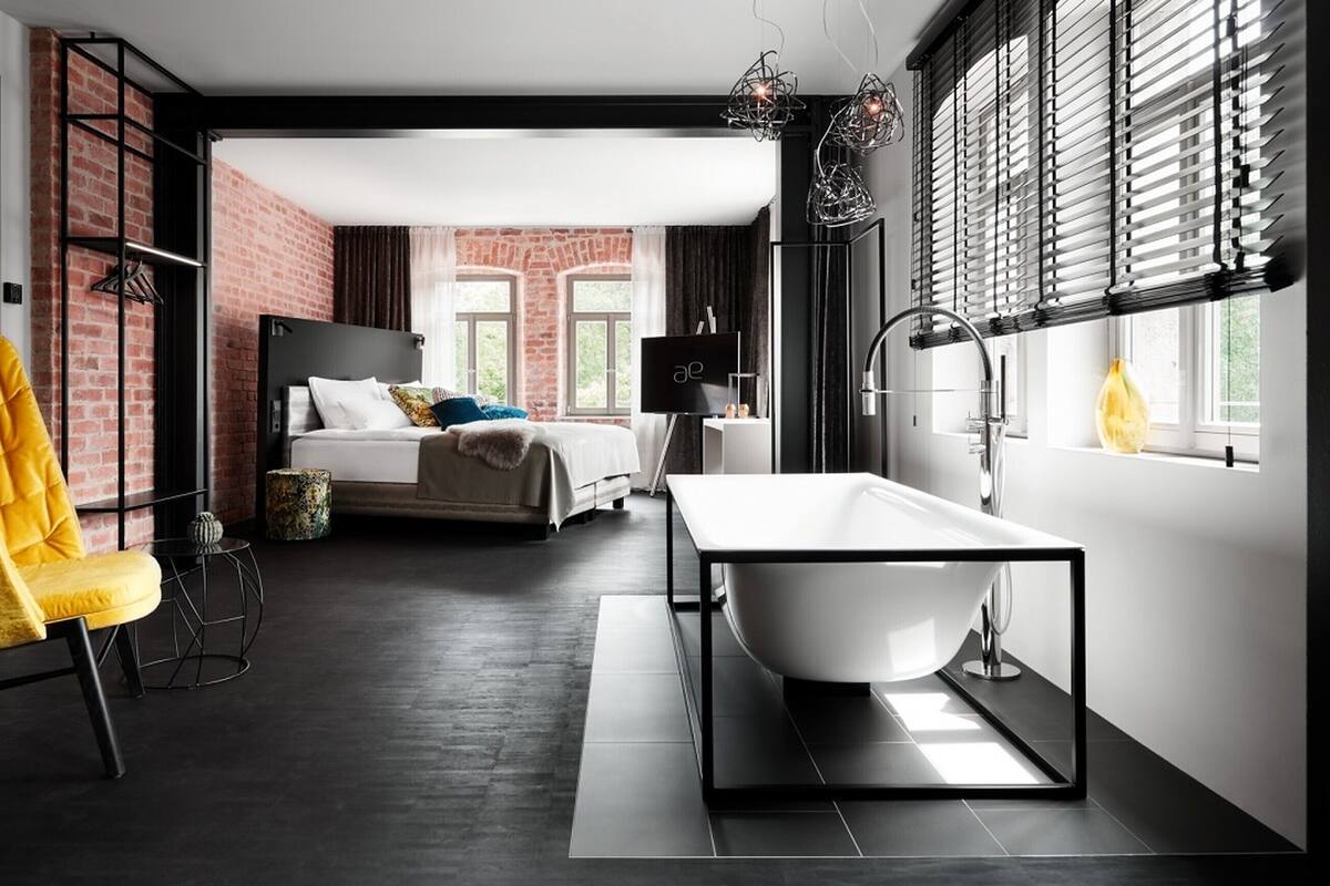 A modern hotel room with a large bed, a yellow armchair, a white bathtub, and a view of a brick wall through the window.