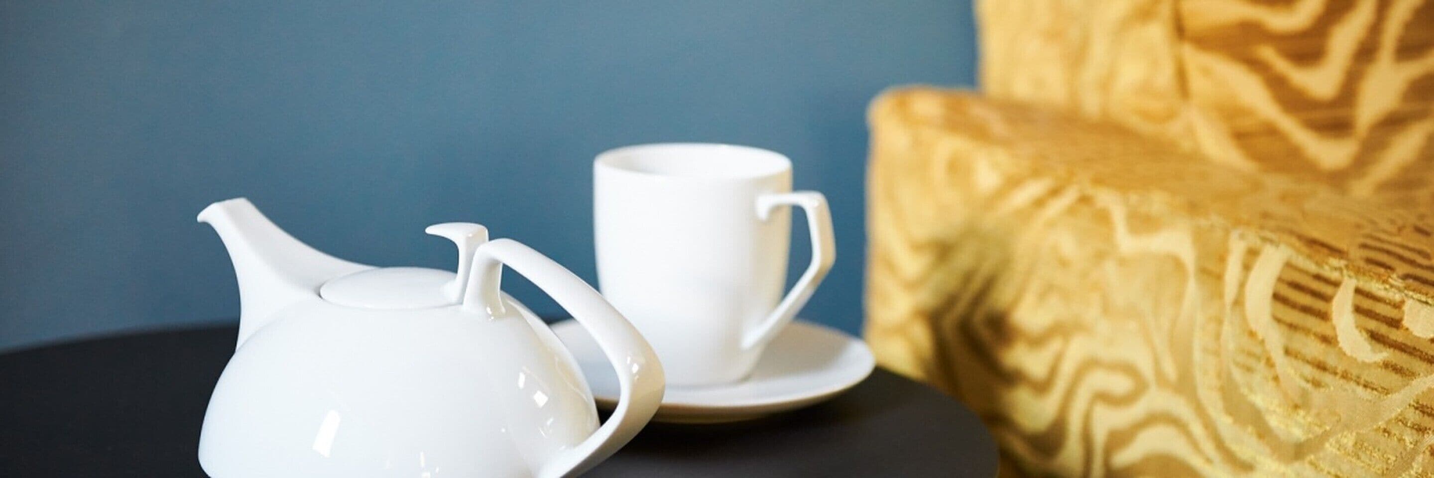 Eine weiße Teekanne und Tasse auf einem schwarzen Tisch, im Hintergrund eine blaue Wand und ein gemustertes Sofa.