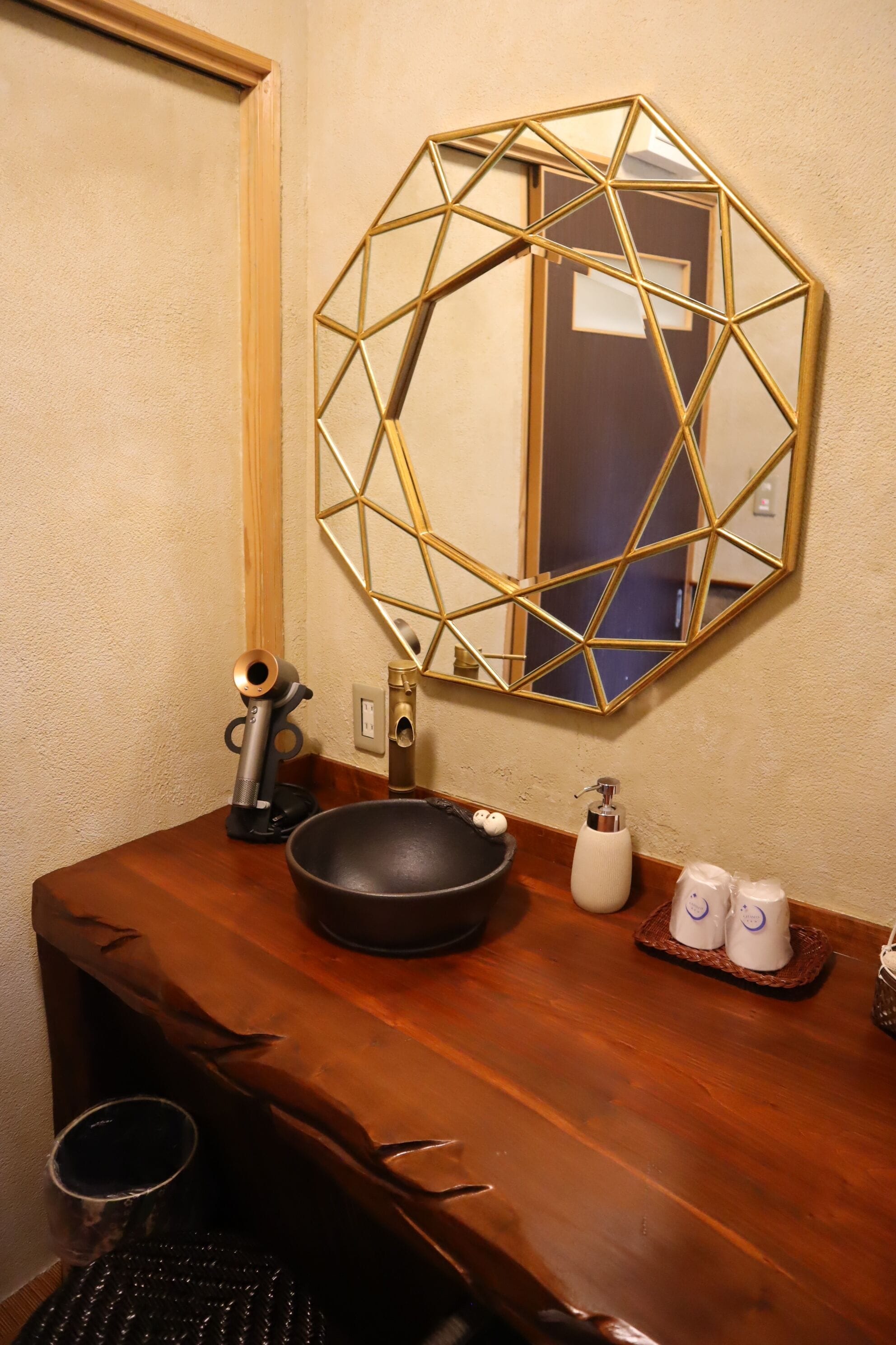 A bathroom with a wooden vanity, a geometric mirror, a black basin, a soap dispenser, and a towel warmer.