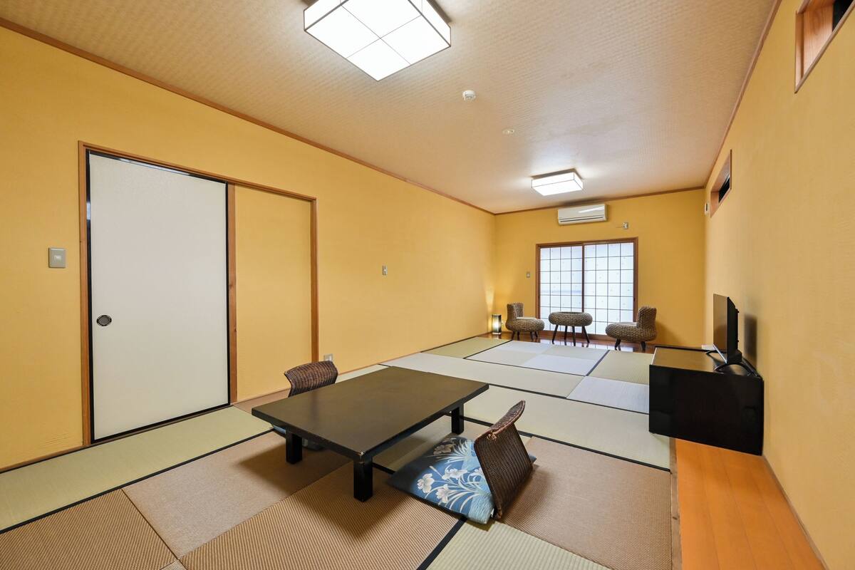 Una habitación con suelo de tatami, una mesa de tatami, sillas, una puerta corredera y un mueble para televisor.