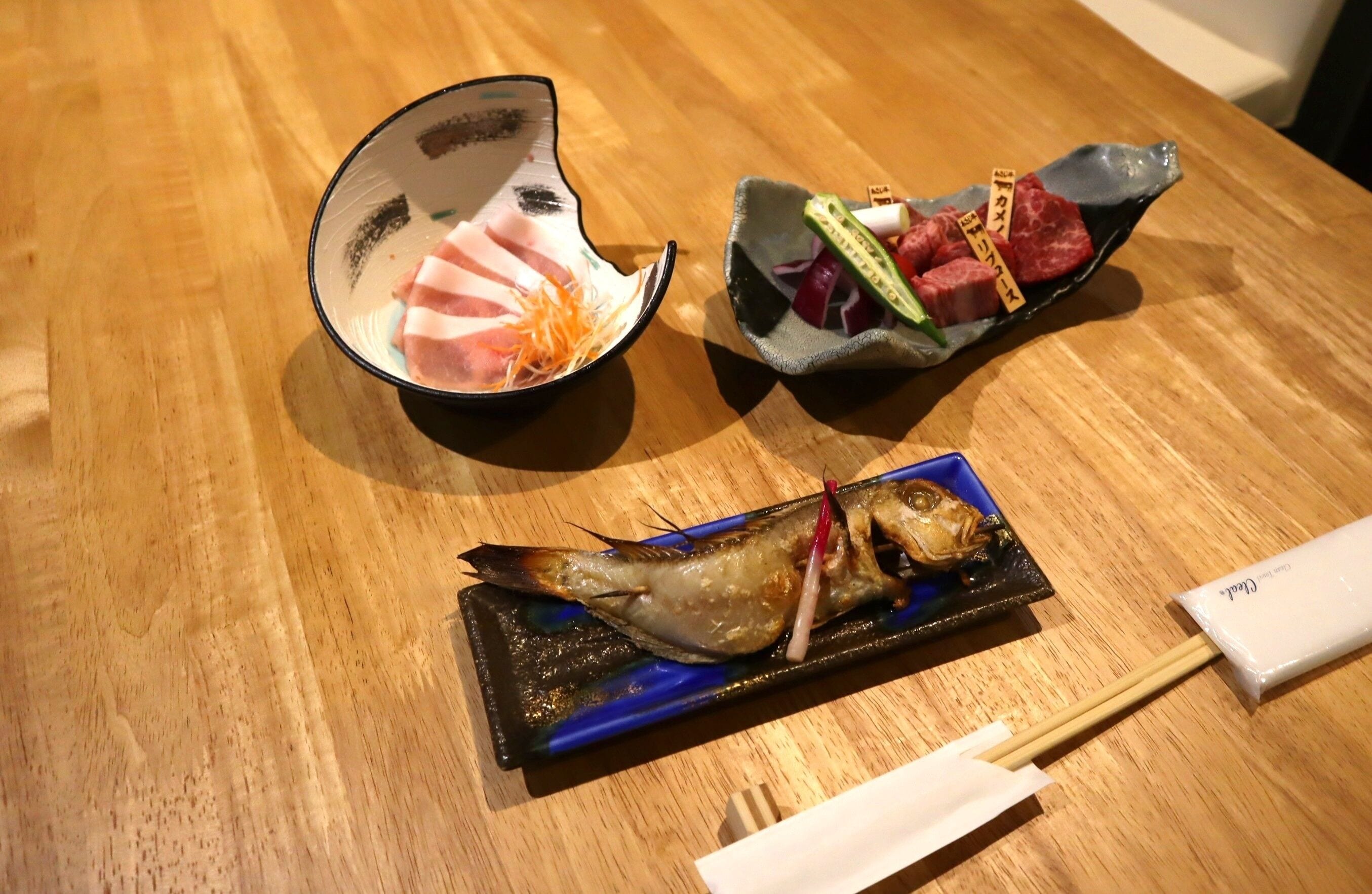 A wooden table with three dishes: a bowl of sliced meat with shredded vegetables, a dish with various meats and vegetables, and a fish on a plate with chopsticks.