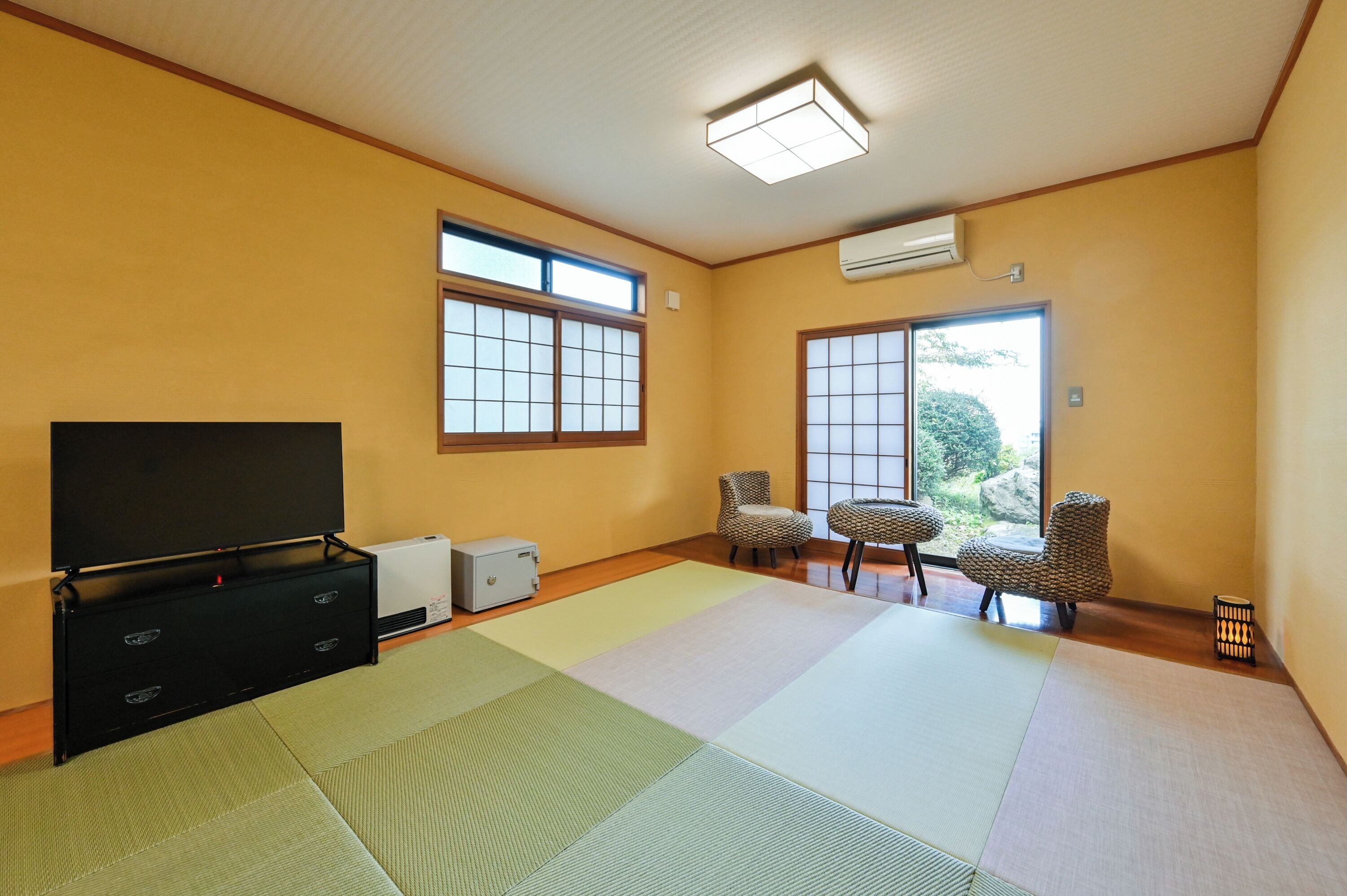 Ein Zimmer mit Tatami-Matten, einem Fernseher, einer Heizung und einem Fenster mit Blick auf Grünflächen.