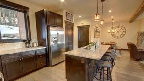 A modern kitchen with dark wood cabinets, a stainless steel refrigerator, a large island with bar stools, and a stone fireplace. - Alders 854 Independence Rd (Keystone)