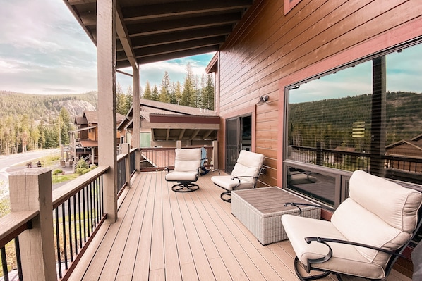 A wooden deck with two white cushioned chairs and a small table, overlooking a forested area.
