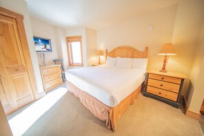 Une chambre à coucher avec un grand lit, une tête de lit en bois, deux tables de chevet avec des lampes, une commode en bois, un téléviseur fixé au mur et une fenêtre avec des stores.