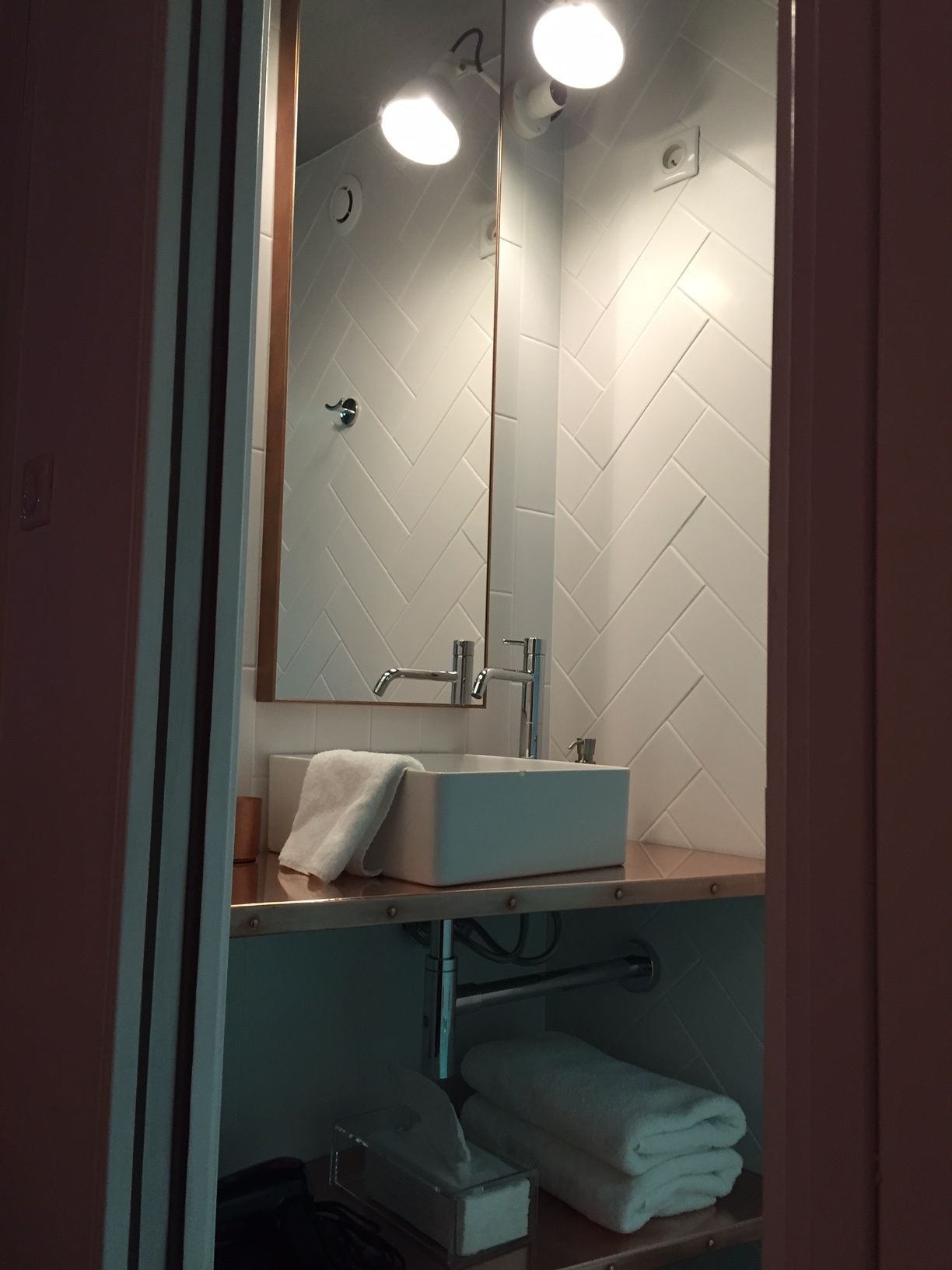 Um banheiro com revestimento de azulejos brancos em padrão herringbone, uma pia retangular e um espelho acima dela. Há uma toalha na pia e um porta-toalhas embaixo.