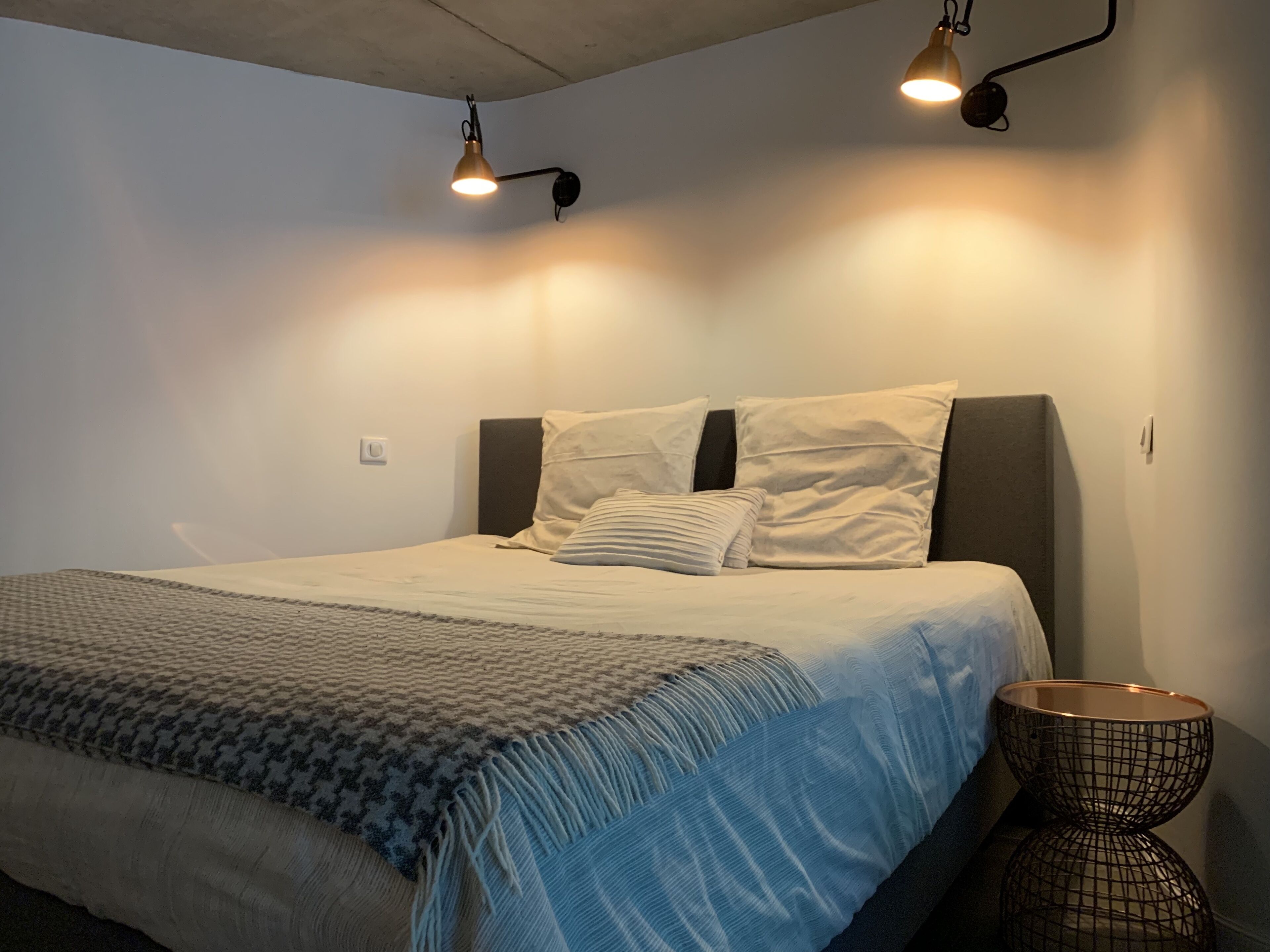 Een net opgemaakt bed met een gebPatronen deken, twee wandlampen en een klein draadmanden aan de zijkant.