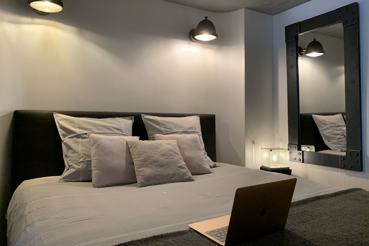 Une chambre à coucher moderne avec un grand lit, un ordinateur portable sur la table de chevet et un grand miroir au mur.